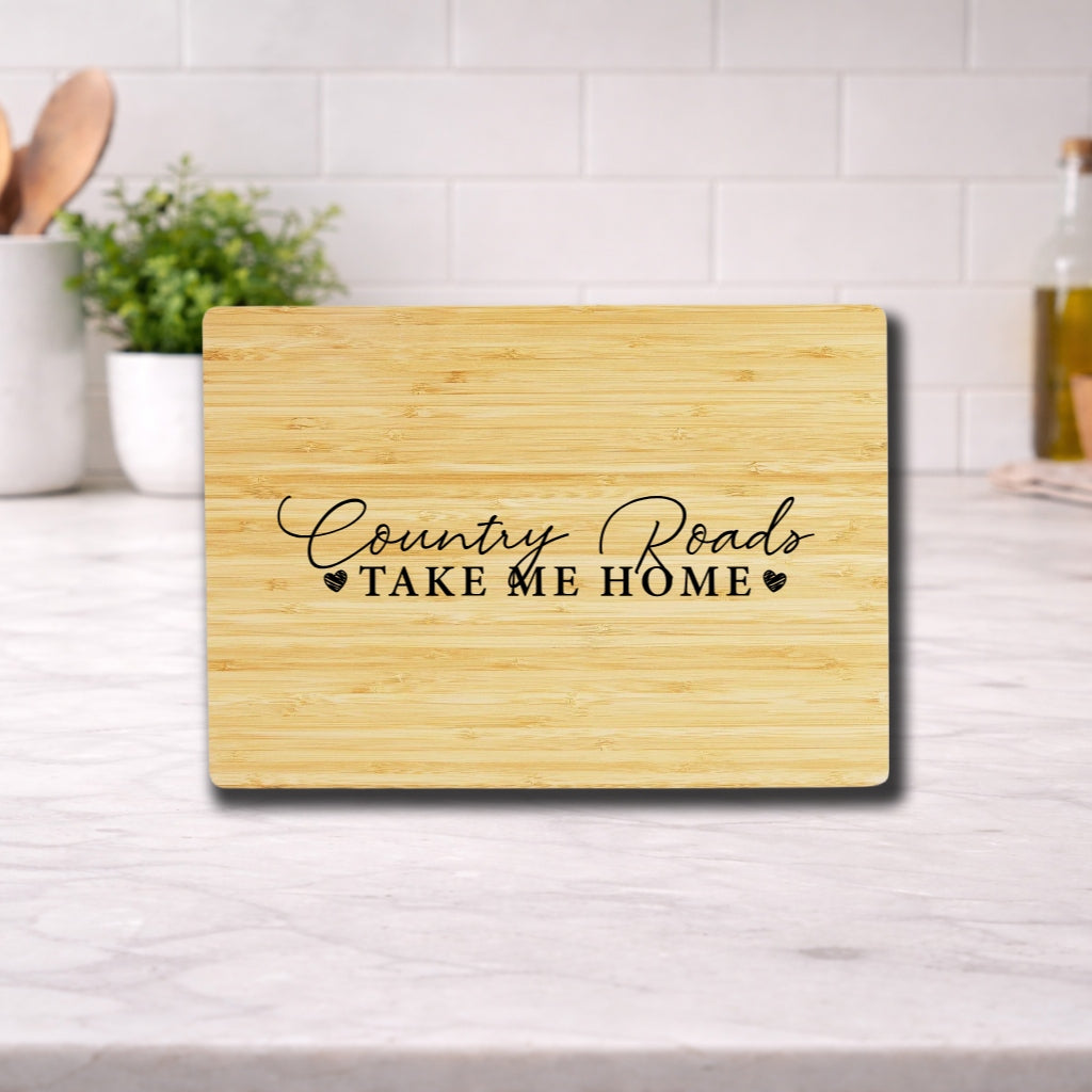 Engraved cutting board with the phrase 'Country Roads Take Me Home' on a wooden surface in a kitchen setting.