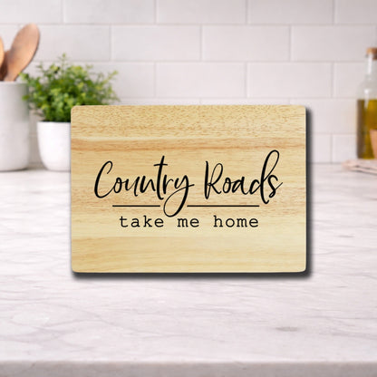 Engraved cutting board with the phrase 'Country Roads Take Me Home' on a wooden surface in a kitchen setting.