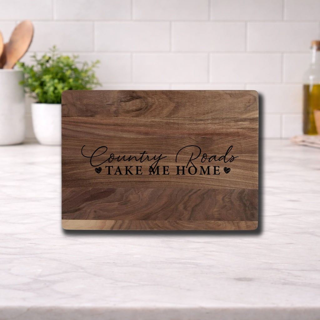 Engraved cutting board with the phrase 'Country Roads Take Me Home' on a wooden surface in a kitchen setting.
