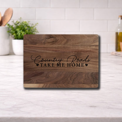 Engraved cutting board with the phrase 'Country Roads Take Me Home' on a wooden surface in a kitchen setting.