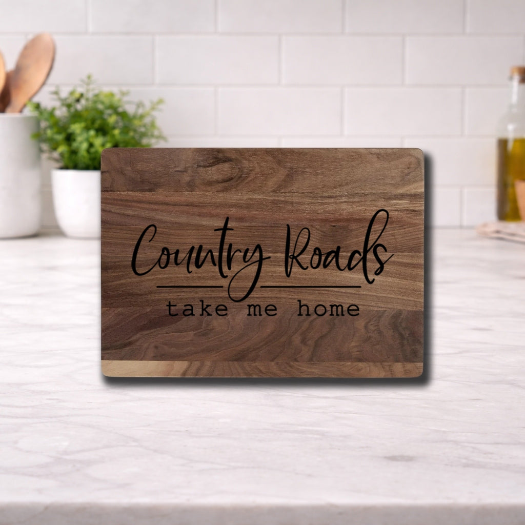Engraved cutting board with the phrase 'Country Roads Take Me Home' on a wooden surface in a kitchen setting.